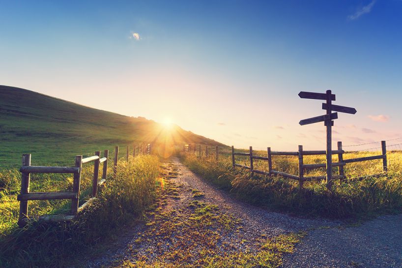Dirt path leading into the hills with setting sun behind the hill and a wooden sign in the foreground pointing three different directions.