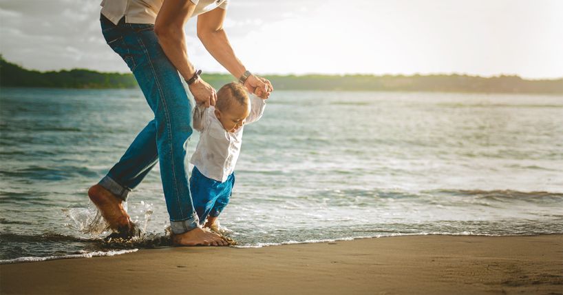 Parent holding hands of very young child to help them walk on the beach.