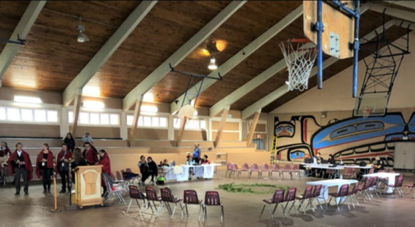 Group of people standing in a line wearing blankets near chairs arranged in a circle in a community hall.